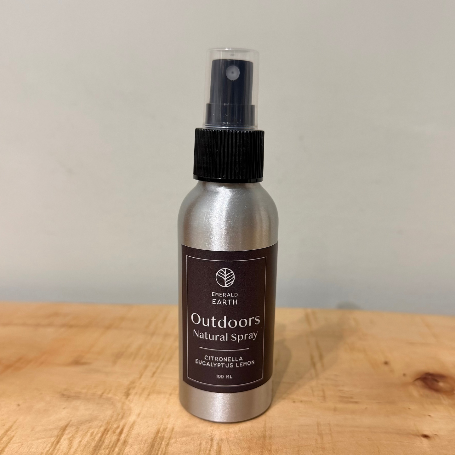 A silver aluminum spray bottle of Outdoors Natural Spray by Emerald Earth, featuring a black spray nozzle and a black label, standing on a light wooden surface against a plain off-white wall.