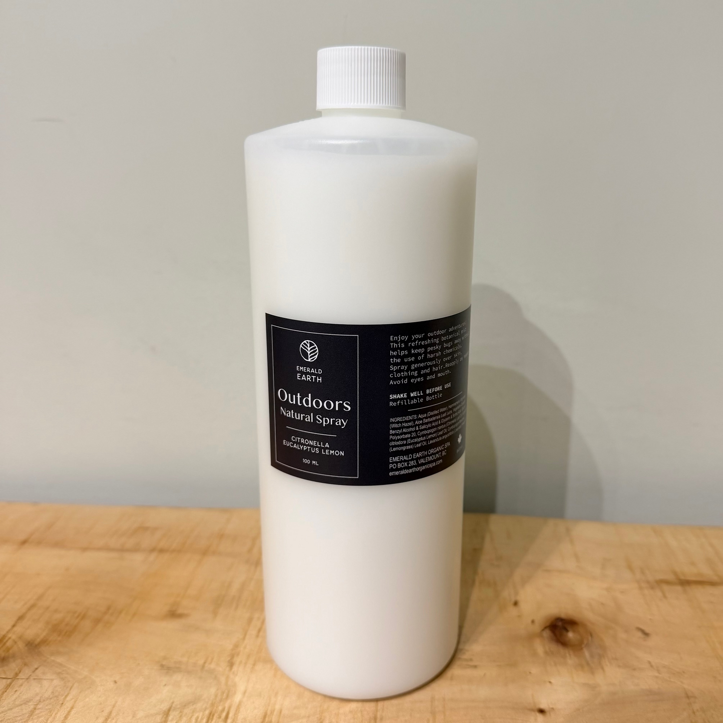 A large white plastic bottle of outdoor Natural Bug Spray standing on a wooden surface against a plain light gray wall.