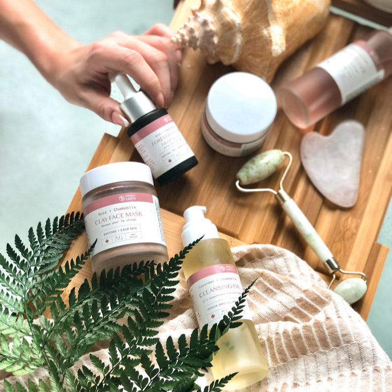 Skincare products on a wooden tray with a hand reaching for a bottle, surrounded by ferns and shells.