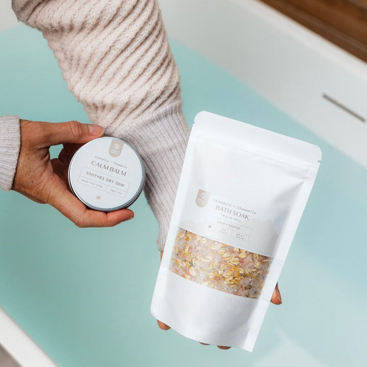 Person holding a body salve and a bag of bath salts product near a bath tub