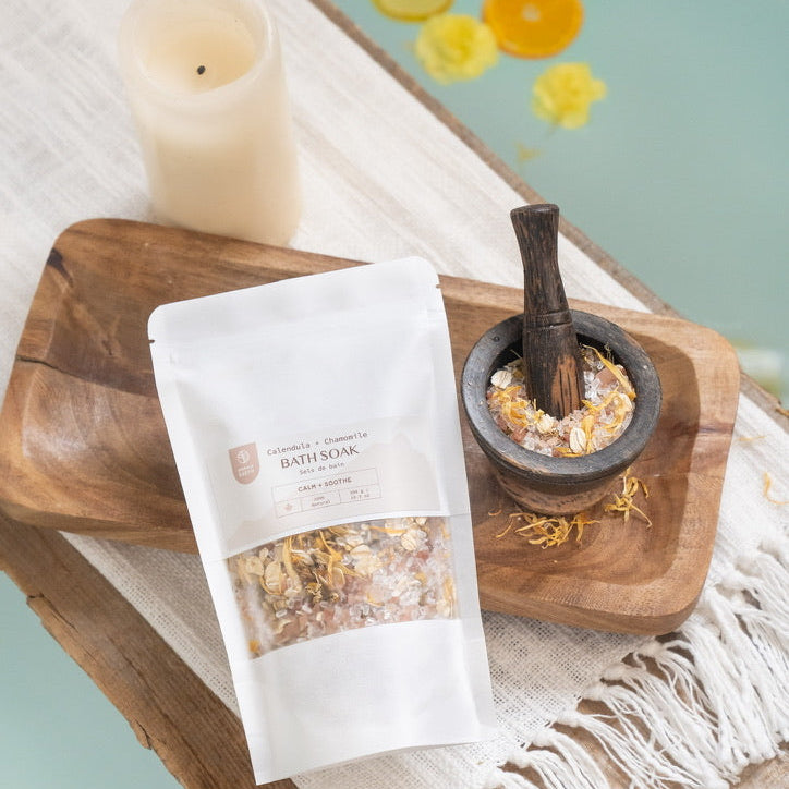 Wooden tray with a bag of bath salts, mortar and pestle, and decorative elements on a light blue background.