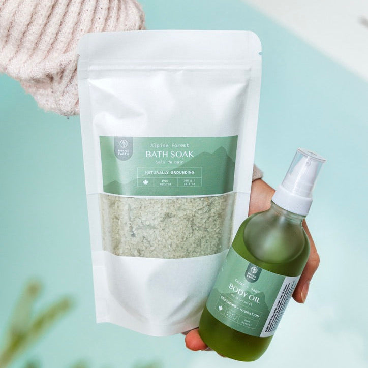 Hand holding a bath soak package and a bottle of body oil on a light green background