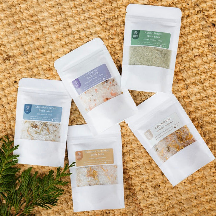 Five white packages of bath soaks on a cork surface with greenery.