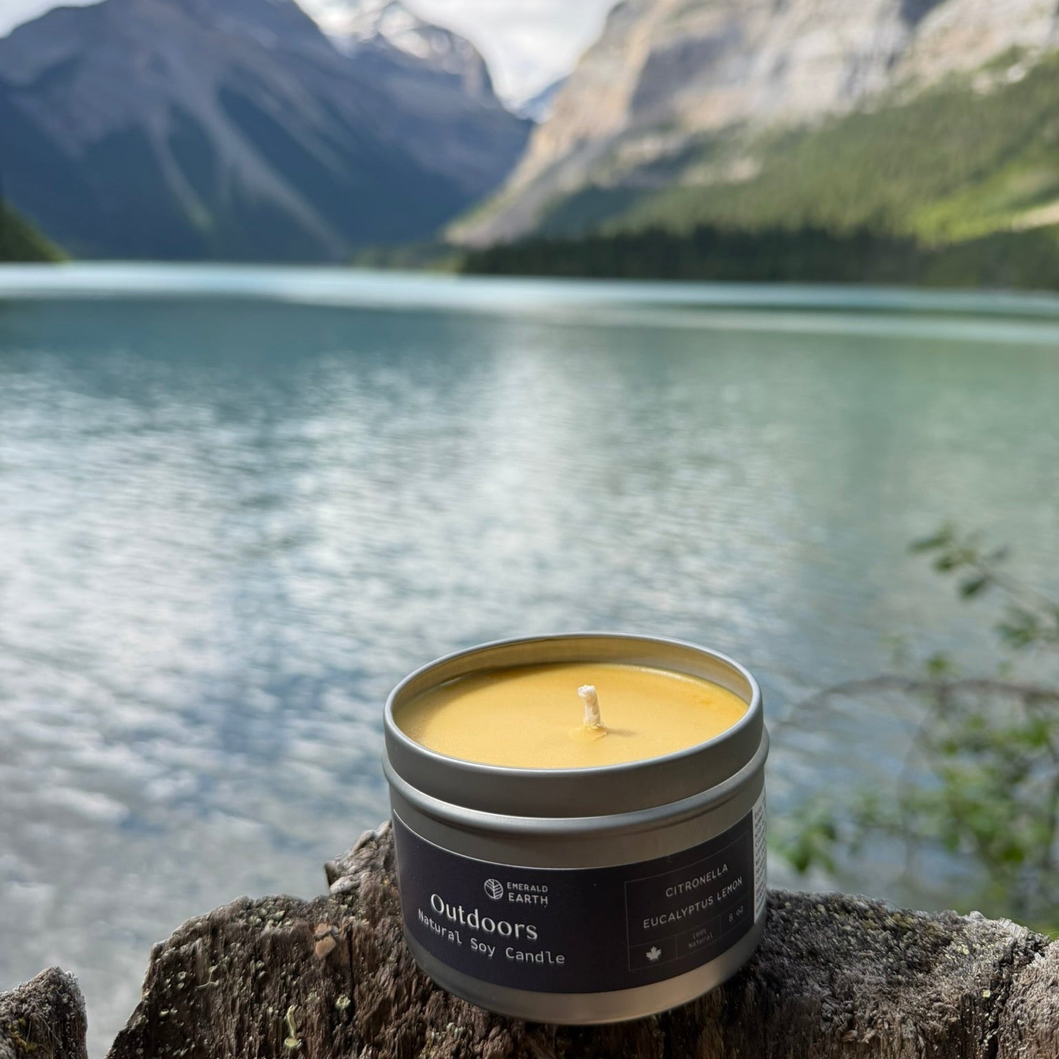 A single-wick soy candle in a silver tin container sits on a weathered tree stump in front of a scenic mountain lake landscape. The candle label reads 'outdoors' and 'Soy Candle'.