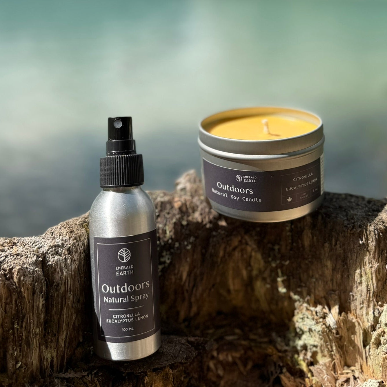 Two outdoor-themed products from the brand Outdoors by Nature: a silver atomizer spray bottle and a silver tin candle, both with black labels, displayed on a rustic tree stump with a blurred mountain and lake background.