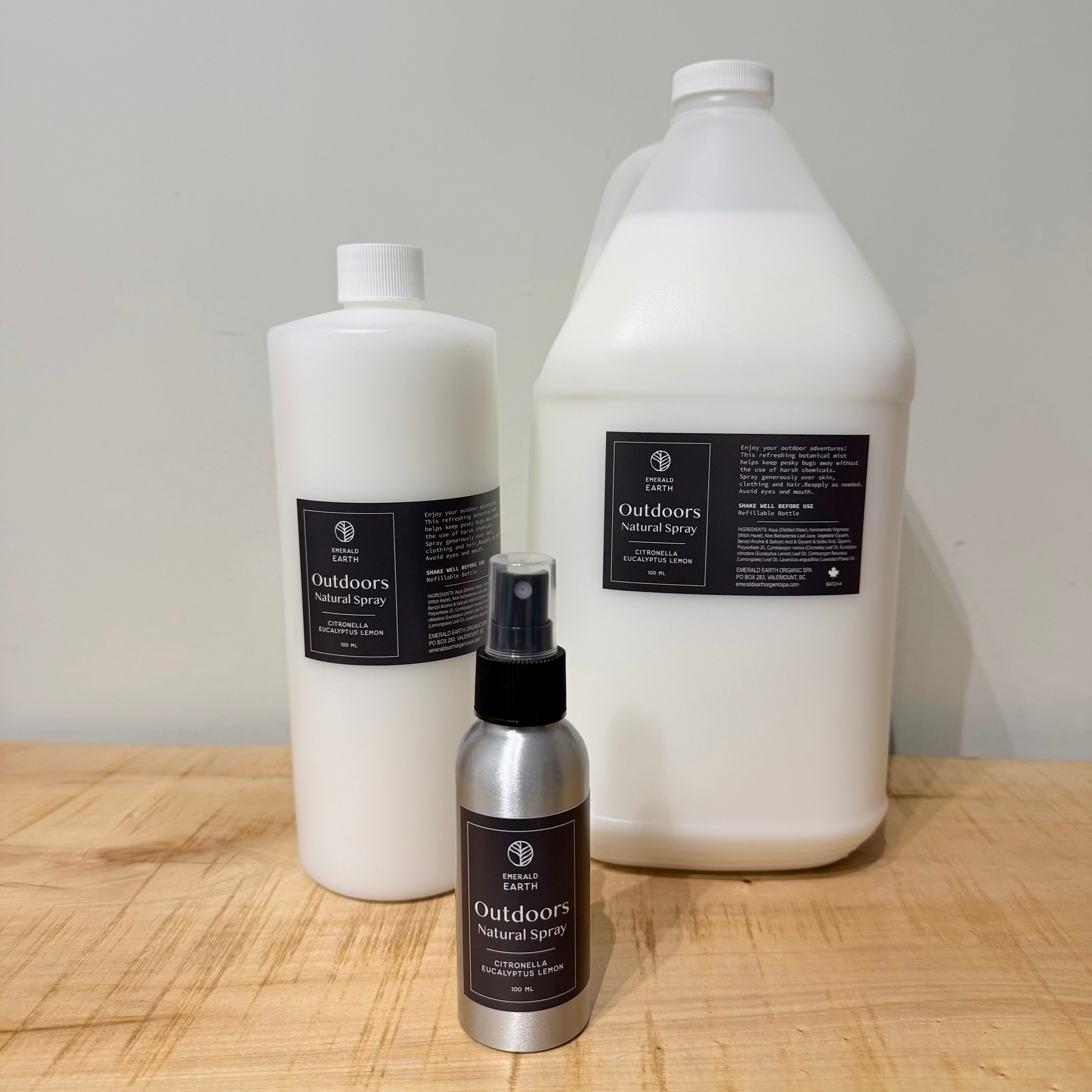 Three bottles of Outdoors Natural Bug Spray displayed on a light wood surface against a plain light gray wall. The set includes a small silver spray bottle in front, a white plastic bottle in the middle, and a larger white plastic jug in the back, each with black product labels.