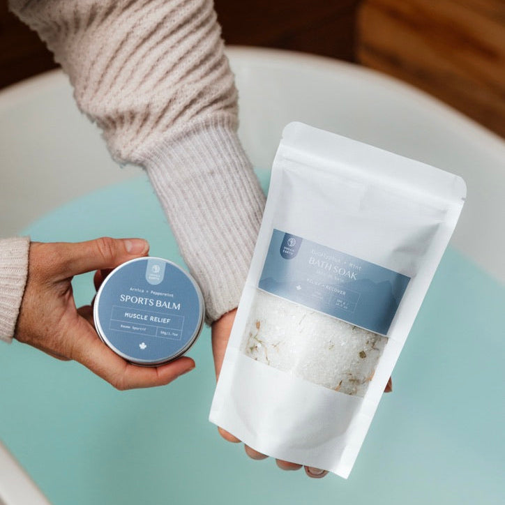 Person holding a jar and a pouch of sports balm on a light blue surface.