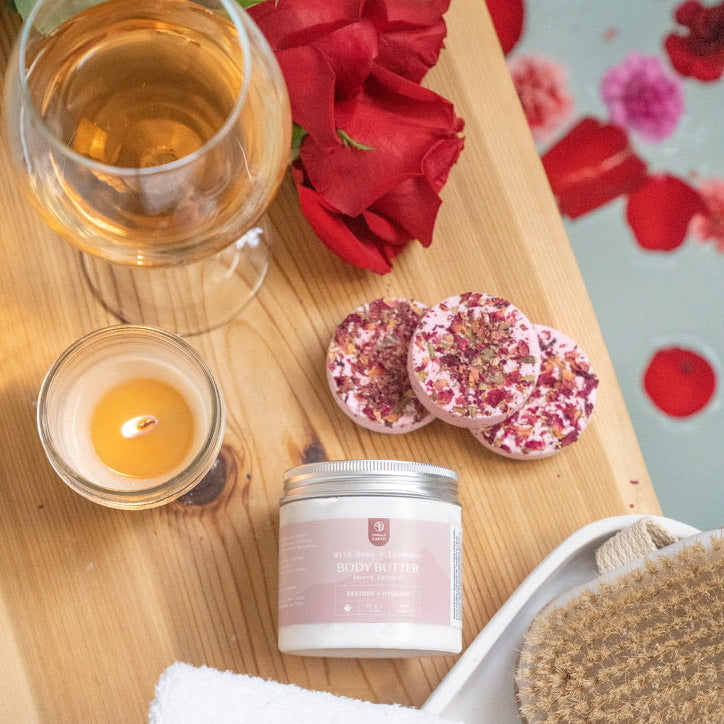 Wooden tray with a glass of white wine, candles, banth bombs, body butter, and a towel on a decorative background with flowers.
