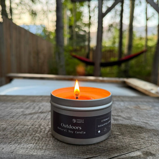 A lit outdoor candle in a silver tin with a dark label reading 'Outdoors Natural Soy Candle' sits on a weathered wooden surface, with a blurred backyard scene including trees and a hammock in the background.