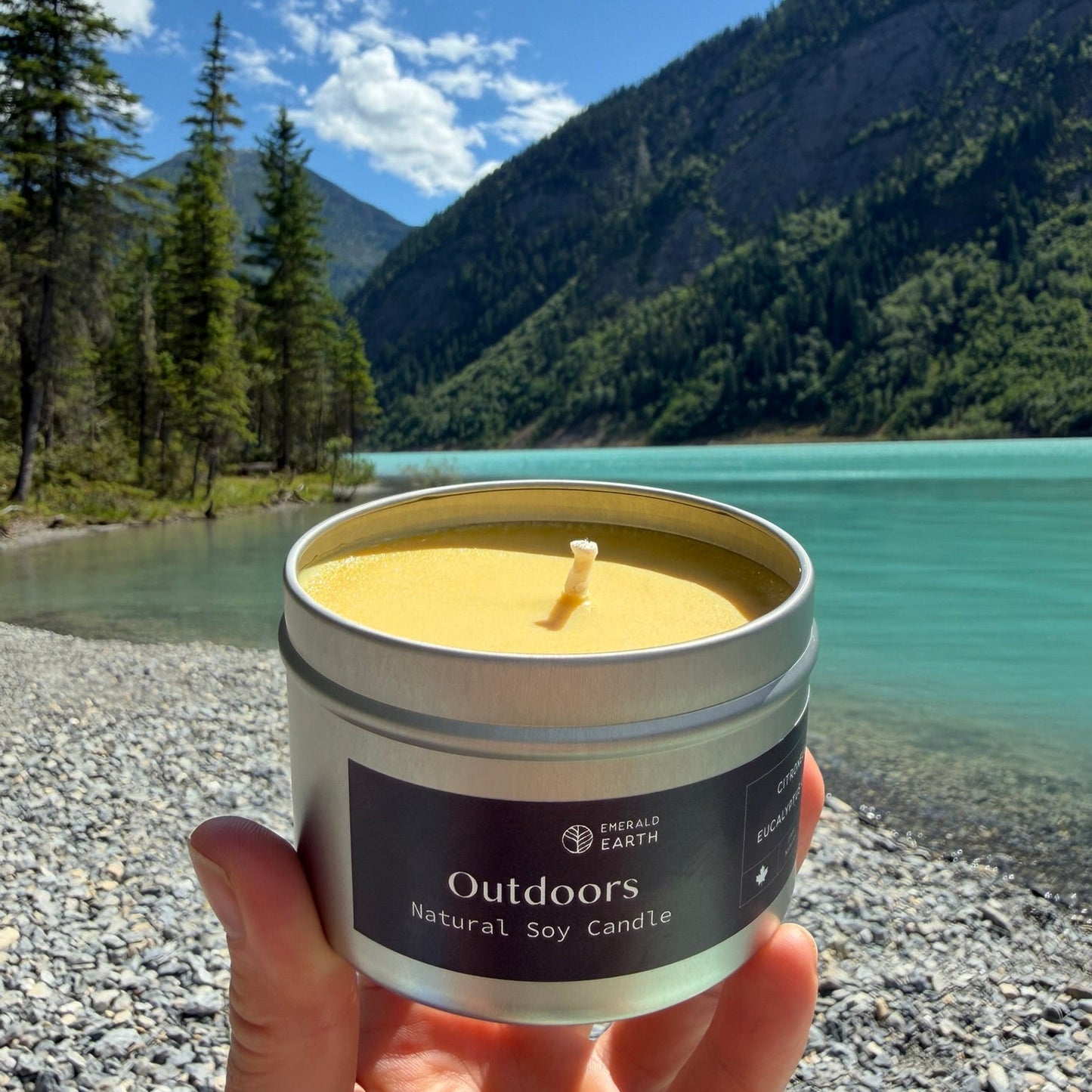 A hand holds a silver tin candle labeled 'Outdoors Natural Soy Candle' in front of a scenic mountain lake landscape.