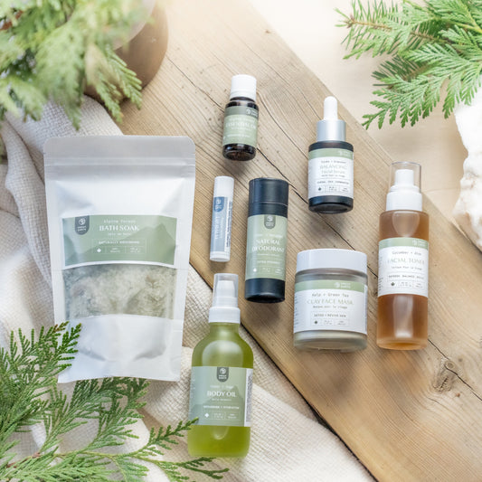 A collection of bath and body products laid out on a wooden surface, including a bath soak, body oil, natural deodorant, essential oil blend, facial serum, facial toner, clay face mask, and lip balm, all with a forest theme in terms of packaging and product names.